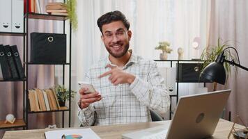 Smiling young businessman using smartphone app online pointing showing like thumbs up remote distance job working from home office. Guy freelancer e-learning texting messages content on social media. video
