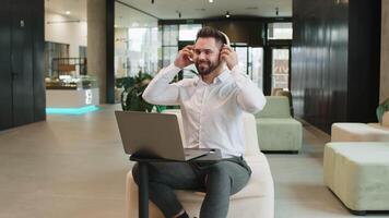 Happy overjoyed Caucasian bearded businessman sitting in modern office lobby listening to energetic disco music through wireless headphones. Manager guy relaxing taking a break with laptop on table. video