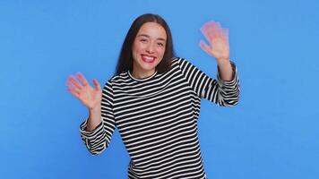 Young Caucasian woman smiling friendly at camera, waving hands gesturing hello greeting or goodbye welcoming with invitation hospitable expression. Pretty girl isolated on blue studio background video