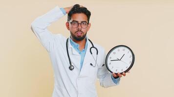 Indian young doctor cardiologist man with anxiety checking time on clock, running late to work, being in delay, deadline. Arabian pharmacy guy worrying to be punctual isolated on beige background video