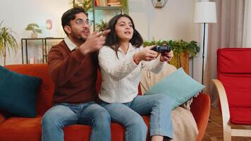 Cheerful young Indian couple using joystick controller playing game fun enjoying sits on sofa in living room. Diverse family enjoying success winning online game during weekend in house indoor video