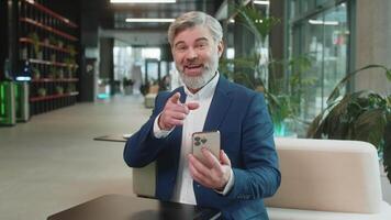 Smiling mature businessman using smartphone app online pointing showing like thumbs up remote distance job working at office lobby. Guy in suit e-learning texting messages content on social media. video