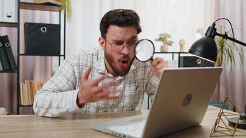Surprised investigator researcher businessman working at home office holding magnifying glass looking at laptop screen with wow expression, searching, analyzing. Professional freelancer at home office video