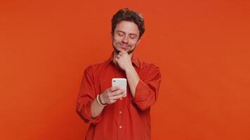 Caucasian man texting share sms messages content on smartphone social media applications online, watching relax movie, browsing. Young guy uses mobile phone smile isolated on orange background indoors video