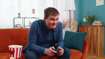 Cheerful Caucasian young man using joystick controller playing game fun enjoying sitting on sofa couch in living room. Guy enjoying success winning online game during weekend at home apartment. video