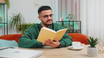 Indian man reading interesting book, turning pages smiling enjoying literature, taking a rest on comfortable couch. Portrait of peaceful cheerful guy relaxing at home apartment living room at table video