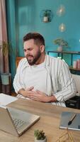 Disappointed freelancer man feeling overeating during work process on laptop in home office. Upset businessman freelancer guy man touching stomach and experiencing discomfort and sharp pain. Vertical video