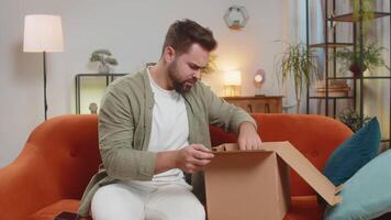 Unhappy young Caucasian man guy unpacking carton box, feeling dissatisfied with getting damaged wireless headphones order, writing negative feedback in mobile application, internet shopping concept. video
