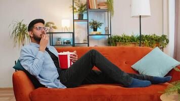 Relaxed Indian young man eating popcorn and watching movie wow expression lying on sofa in living room at home. Happy Hispanic guy in casual clothes enjoying film during weekend in apartment room. video
