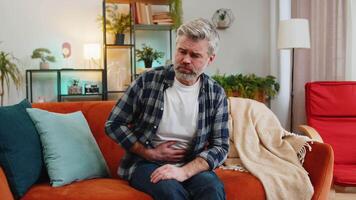 Mature ill sick man sitting on couch feeling sudden strong abdominal stomach ache, gastritis problem. Middle-aged guy having symptom poisoning diarrhea indigestion peptic ulcer pancreatitis at home. video