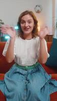 Happy young Caucasian woman smiling friendly at camera and waving hands gesturing hello, hi, greeting talking while sitting on sofa in living room at home. Happy girl recording vlog at home. Vertical video