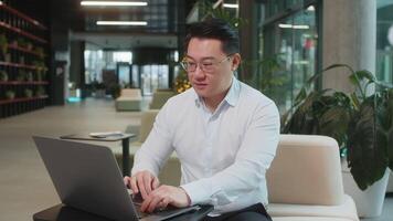 kinesisk ung affärsman stängning bärbar dator dator efter efterbehandling arbete på hotell lobby. leende chef upptagen arbetssätt uppkopplad, avlägsen jobb. e-lärande, bläddring internet på netbook ser på kamera. video