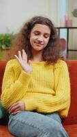 Young child girl smiling friendly at camera and waving hands gesturing hello, hi, greeting or goodbye, welcoming with hospitable expression. Teenager kid at home in living room sits on sofa. Vertical video