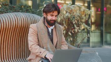 Indian business man freelancer closes laptop screen finishing working online distant job browsing website chatting, looking at camera. Bearded guy use notebook, sends messages on downtown city street video