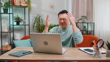 Displeased sad Asian man use laptop notebook typing browsing working, loses becoming surprised sudden lottery results bad news fortune loss game fail computer virus. Chinese guy at home room on couch video