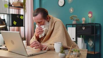 Sick ill senior man suffering from cold flu or allergy working on laptop at home office. Manager freelancer grandfather wrapped in blanket blows sneezes wipes snot into napkin takes medicine at table. video