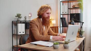 Caucasian bearded businessman working on laptop shocked by sudden victory, game winning lottery goal achievement, good career news. Excited redhead guy freelancer clenching fists at home office. video