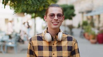 Portrait of happy man with headphones around neck smiling friendly, glad expression looking at camera dreaming, resting, relaxation feel satisfied good news outdoors. Young guy in urban city street. video