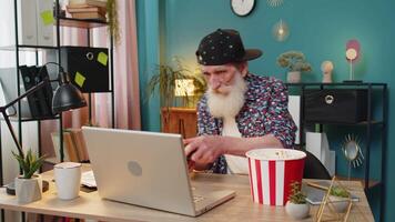 Excited Caucasian senior man celebrating victory in game on laptop clenching fists, spending leisure time at home office desk. Happy stylish grandpa playing games holding controller. video