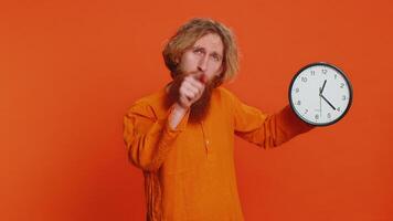 It is your time, hurry up. Caucasian man showing time on wall office clock, ok, thumb up, approve, pointing finger at camera, advertisement, deadline. Redhead young guy isolated on orange background video