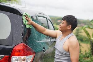 Asian man washing his car with care using a green microfiber cloth. The scene shows a maintenance routine done at home, promoting responsibility, cleanliness, and pride in vehicle ownership. photo