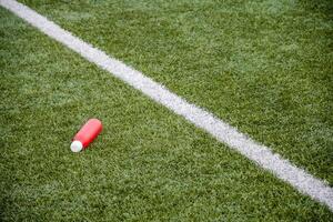 red water bottle lies on the green artificial grass of a football field near the dividing white line, the concept of drinking during long workouts and replenishing fluids and electrolytes, photo