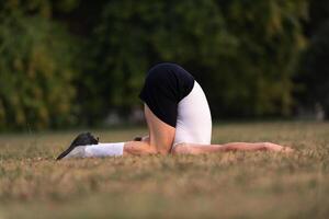 A fit man lying on the grass in a yoga or stretching pose, with head tucked under and legs bent, in a natural park setting. photo