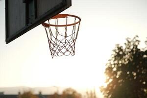 A basketball hoop at an outdoor court captured in the warm light of sunset. The minimalist and nostalgic scene highlights the hoop and net against the silhouette of trees and sky. photo
