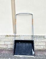 Black snow shovel with metal handle against wall of building and cleaned sidewalk. Winter weather, snow removal, cleaning roads and driveways photo