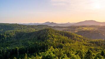 A stunning panoramic view from Popov Cliffs reveals the lush green hills and sandstone formations of the Lusatian Mountains during a tranquil evening at sunset. photo