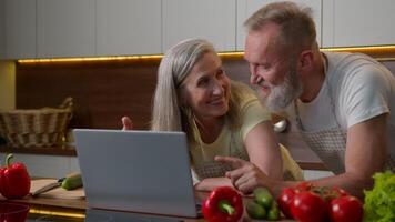 Caucasian family middle-aged married couple woman and man use laptop online shopping buying fresh food internet store pay order retired husband wife laughing talking old age and technology in kitchen video