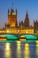 Westminster Palace stands illuminated against the twilight sky, reflecting on the Thames River. The glow of the buildings and bridge adds charm to this iconic location at dusk. photo