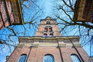 Visitors admire the stunning architecture of the Church of Our Saviour in Copenhagen, Denmark, framed by bare branches, showcasing the tower against a bright blue sky. photo