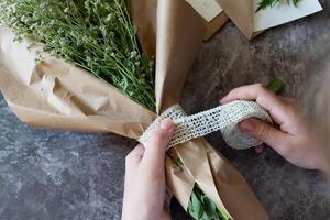 a woman prepares a summer bouquet of white gypsophila on a gray desk. Kraft paper, scissors, greeting envelope on the table. View from above. The composition is flat. photo