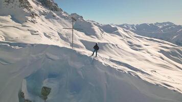 Aerial circular revealing view man skier traveler or tourist stands at Tetnuldi snowy mountain summit viewpoint next to cross in bright winter landscape Caucasus Georgia inspirational scenery video