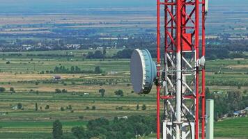 A red and white telecommunication tower with a large dish antenna stands in a rural area with green fields, scattered trees, and distant villages at summer day. video
