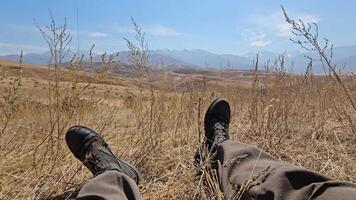 First-person perspective POV shot of legs in military combat boots resting in dry grass, enjoying the mountain landscape view in Kyrgyzstan. video