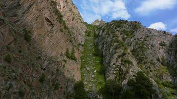 Aerial footage capturing a flight along a rugged mountain chute in Kyrgyzstan, following a natural rocky path through steep cliffs and sparse greenery under a clear blue sky. video
