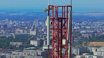 A telecom tower with panel and dish antennas stands tall above a dense urban Bishkek cityscape, featuring high-rise buildings, construction sites, and residential areas. Drone view, arc movement. video
