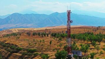 Drone footage of a telecom tower in countryside, surrounded by dry hills and scattered trees. The drone moves slowly in an arc around the tower, showing parallax with distant mountains in the background. video