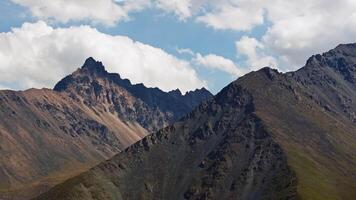 en Drönare fångar en fantastisk antenn se av en oländig berg räckvidd i Kirgizistan, flygande vänster med en lastbil till de vänster kamera rörelse, skapande en dynamisk parallax effekt under en delvis molnig himmel. video