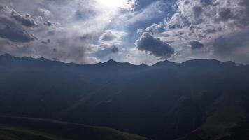 Aerial hyperlapse capturing the rugged mountains of Kyrgyzstan, shot with a drone moving forward. Dramatic clouds cast shadows over the peaks, with sunlight breaking through, highlighting the serene, untouched terrain. video