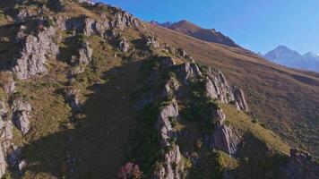 Aerial shot of a mountain slope with rocky cliffs in Kyrgyzstan, camera moving right showing distant snow-covered peaks on the horizon. Sunny evening, clear sky. video
