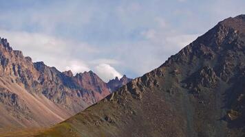 A drone with telephoto lens captures a sweeping arc-left view of rugged mountains in Kyrgyzstan under a partly cloudy sky. The scene showcases rocky peaks, steep slopes, and green valleys with dramatic shadows. video