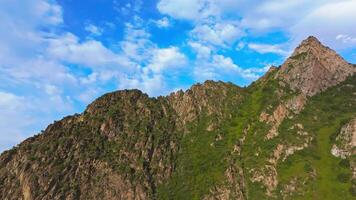 Aerial shot of a rugged mountain formation in Kyrgyzstan at sunset. The camera moves in a circular arc to the left, revealing the volume of the steep, rocky slope with green patches under warm, golden light. video
