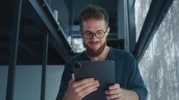 Bearded IT Specialist wearing glasses and casual clothes, smiling and using digital tablet in modern designed office space with large windows. Software Developer develops new mobile application video