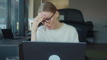 Financial analyst woman suffering from headache while working on laptop in modern office, touching her forehead with hand, showing tiredness and frustration. Fatigue at work in office video