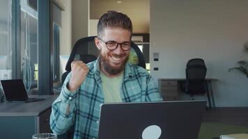 Happy businessman raising fist and smiling while looking at laptop screen, celebrating successful project or deal in modern office workspace. Concept successful transaction or winning video