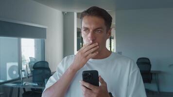 Shocked businessman reacting to bad news on his smartphone while standing in a sleek, modern office, displaying a mix of disbelief and concern on his face amidst the corporate environment video