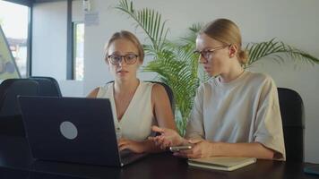 Two young businesswomen online tutor at a modern office table, discussing strategies and working on laptop, exemplifying teamwork and professionalism. Online tutor office workers video